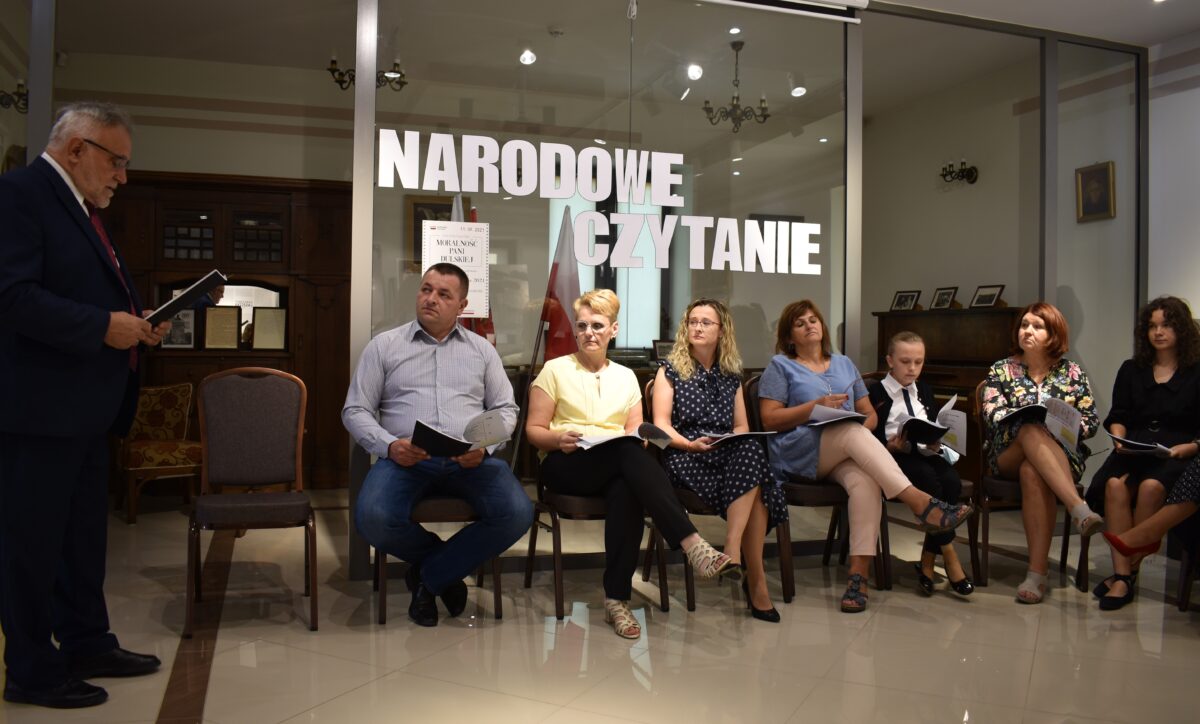 Pan Wójt czyta, reszta uczestników siedzi z zaciekawieniem słuchając. W tle eksponaty Muzeum Konstantego Laszczki