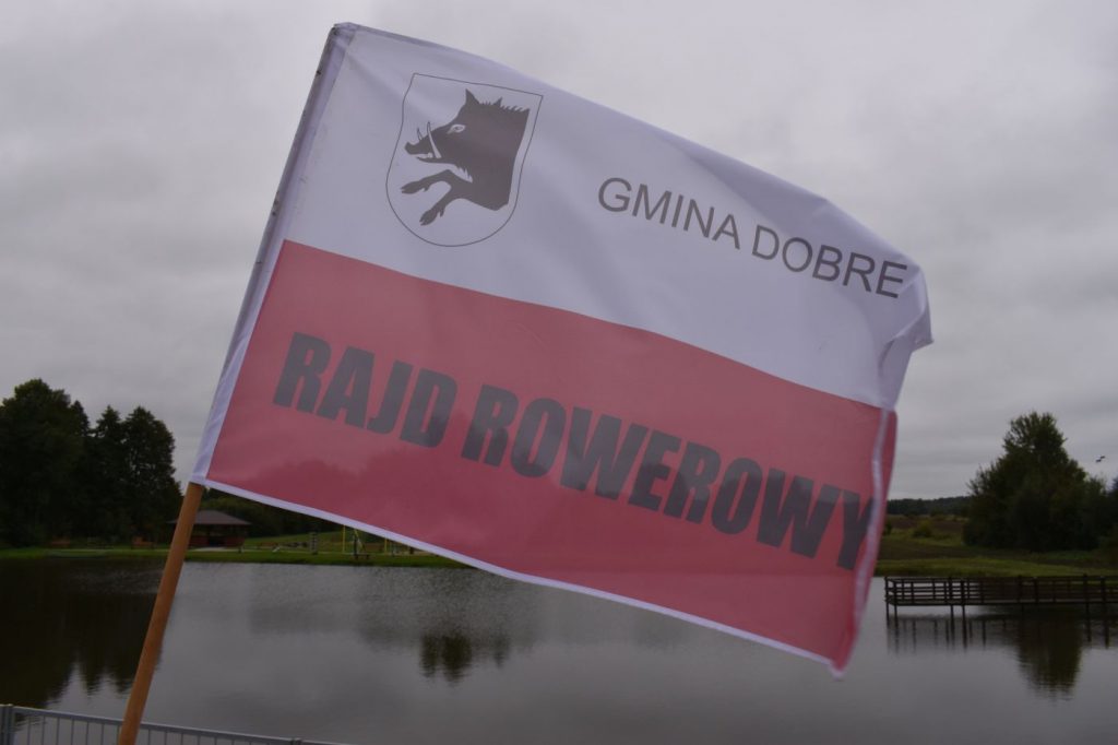 Flaga biało – czerwona na której jest herb przedstawiający głowę dzika i napis Gmina Dobre Rajd Rowerowy, w tle staw oraz drzewa.