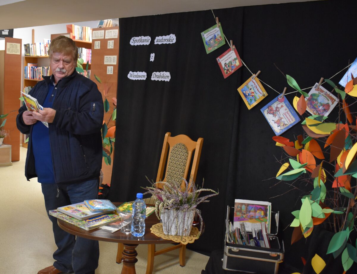 Mężczyzna stojący obok stolika na którym leżą książki, jedną z nich trzyma w ręku. Za nim czarna kotara na której wiszą książki, oraz napis spotkanie autorskie z Wiesławem Drabikiem. W tle widać regały z książkami.