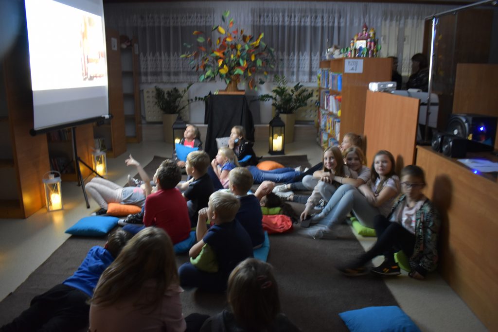 W sali przed ekranem na kolorowych poduszkach siedzą dziewczynki i chłopcy. W oddali widać świecące lampiony i kwiaty