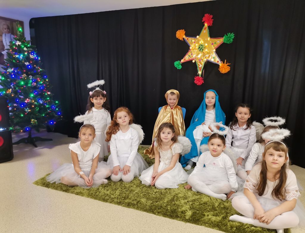 Grupa dzieci siedząca na dywanie ubrana na biało, choinka z kolorowymi światełkami stoi z boku. Na czarnym tle kolorowa gwiazda