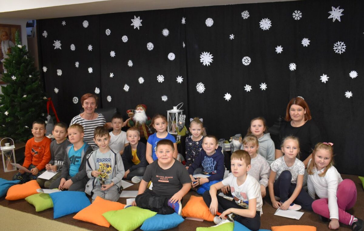 Uśmiechnięta grupka przedszkolaków siedzi na dywanie wraz z dwiema paniami. Przed nimi leżą kolorowe poduszki, za nimi stoi choinka i wiszą białe gwiazdki.