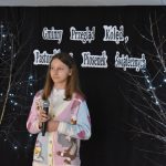 13. Na tle czarnej kotary stoi dziewczyna w kolorowym sweterku trzyma mikrofon w tle czarna kotara, regał z książkami, drzewka z lampkami