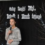 15. Dziewczynka w białej bluzce w czarne paski trzyma mikrofon w ręku na tle czarna kotara z napisem Gminny Przegląd Kolęd, Pastorałek i Piosenek Świątecznych i gałązki ze światełkami
