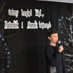 18. Stojący pomiędzy drzewkami chłopiec trzymający mikrofon w reku ubrany w ciemną bluzę w tle czarna kotara z napisem Gminny Przegląd Kolęd, Pastorałek i Piosenek Świątecznych i drzewka ze światełkami