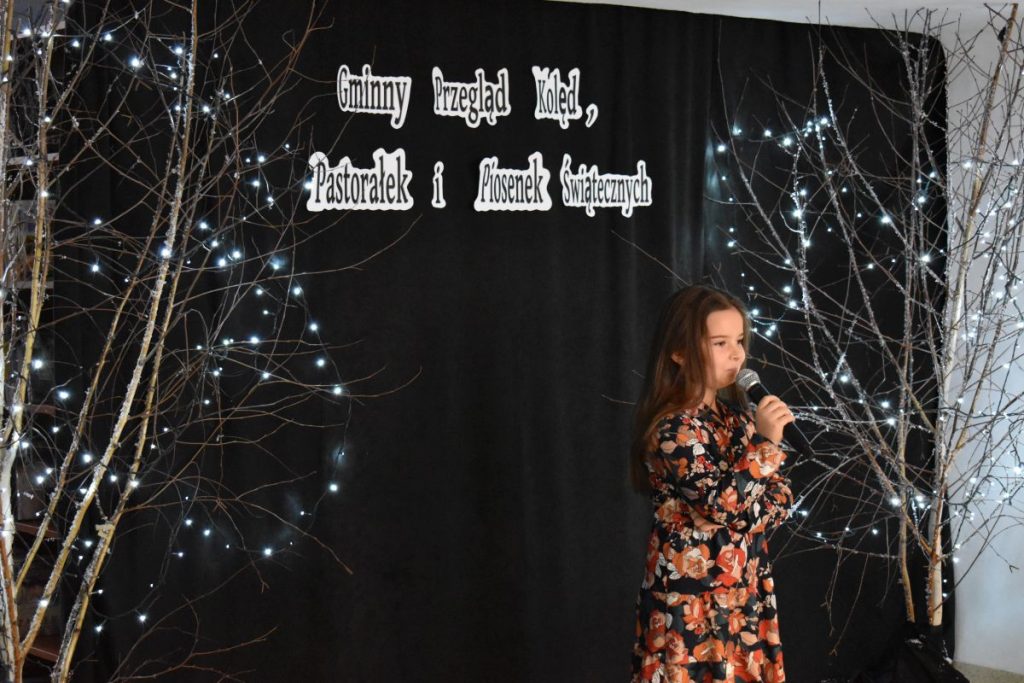 20. Pomiędzy drzewkami ozdobionymi białymi lampkami śpiewa do mikrofonu mała dziewczynka w sukience w kwiaty w tle czarna kotara na której widnieje napis Gminny Przegląd Kolęd, Pastorałek i Piosenek Świątecznych