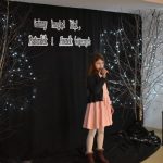 21. Dziewczynka w jasnej sukience i czarnym sweterku trzyma w ręku mikrofon po obu stronach stoję brzozy ozdobione lampkami w tle czarna kotara na podłodze stoi świecący się lampion