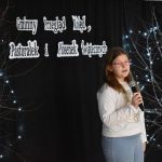 22. Dziewczynka w okularach, białej bluzce i jasnych spodniach trzyma mikrofon w ręku w tle czarna kotara i gałązki ze światełkami oraz napis Gminny Przegląd Kolęd, Pastorałek i Piosenek Świątecznych