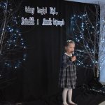 27. Mała dziewczynka z kokardka na włosach w sukience w kratkę z białym kołnierzykiem śpiewa do mikrofonu który trzyma w ręku. W tle czarny materiał na którym wisi napis Gminny Przegląd Kolęd, Pastorałek i Piosenek Świątecznych i dwa drzewka ozdobione lampkami, na podłodze stoją dwa lampiony.