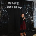 28. Na tle czarnej zasłony z drzewkami ozdobionymi światełkami stoi mała dziewczynka trzyma w raczkach mikrofon, ma na sobie bordową sukienkę i czarny sweterek po prawej stronie stoi latarenka z zapalonymi świeczkami