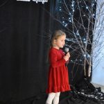 29. W czerwonej sukience i białych rajstopkach stoi mała dziewczynka w rekach trzyma mikrofon, w tle czarna kotara i brzózka ozdobiona lampkami