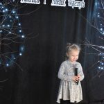 30. Przedszkolaczek w szarej sukience w kwiatuszki trzyma mikrofon, w tle czarna zasłona oraz napis Gminny Przegląd Kolęd, Pastorałek i Piosenek Świątecznych, gałązki i światełka