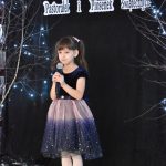 31. Dziewczynka w kucykach trzyma w reku mikrofon, jest ubrana w granatową bluzkę i błyszcząca spódnicę. Na czarnym tle gałązki ze światełkami.