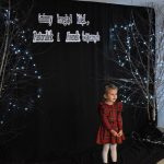 33. Mała dziewczynka w czerwonej sukience w kratkę, stoi pomiędzy dwoma drzewkami ozdobionymi światełkami na podłodze stoi lampion ze świecami w tle czarna zasłona z napisem Gminny Przegląd Kolęd, Pastorałek i Piosenek Świątecznych