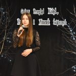 5. Młoda kobieta z długimi włosami w czarnej sukience z mikrofonem w reku. W tle czarna kotara i półki z książka, oraz brzozy z błyszczącymi światełkami