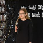 6. Dziewczyna w okularach i czarnej sukience stoi z mikrofonem w dłoni. W tle czarna kotara, półki z książkami, oraz brzozy z błyszczącymi światełkami