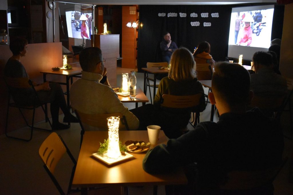 Przy małych stolikach na których stoją lampki, siedzi kilka osób słuchają opowiadającego mężczyzny siedzącego przy dużym ekranie w pomieszczeniu panuje półmrok
