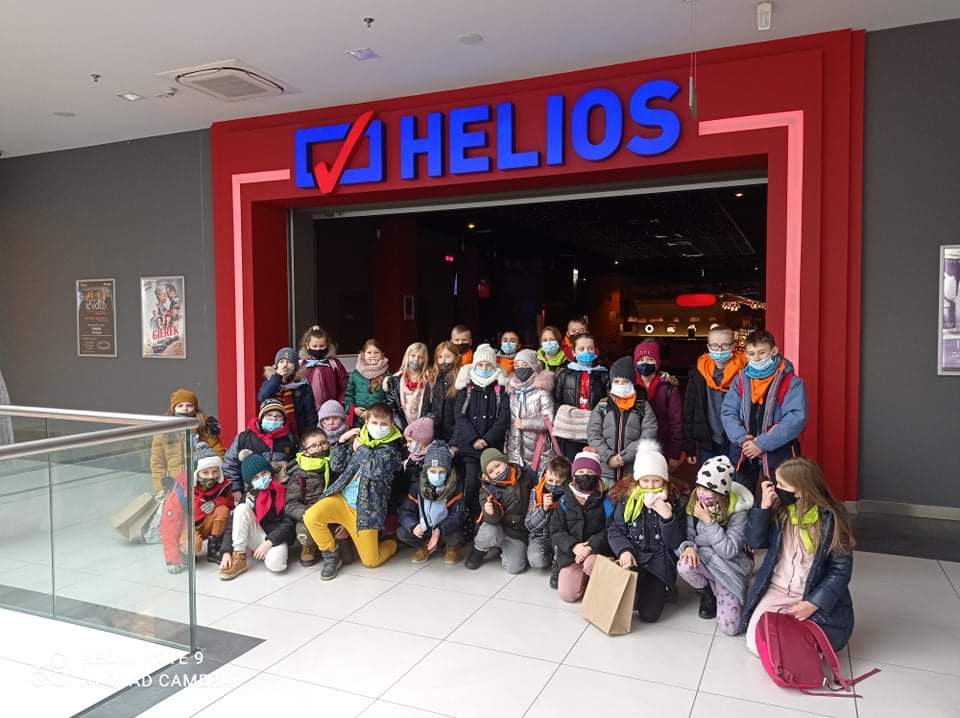 Grupka dzieci część stoi a część kuca. W tle czerwona rama wejścia do kina, z niebieskim napisem HELIOS