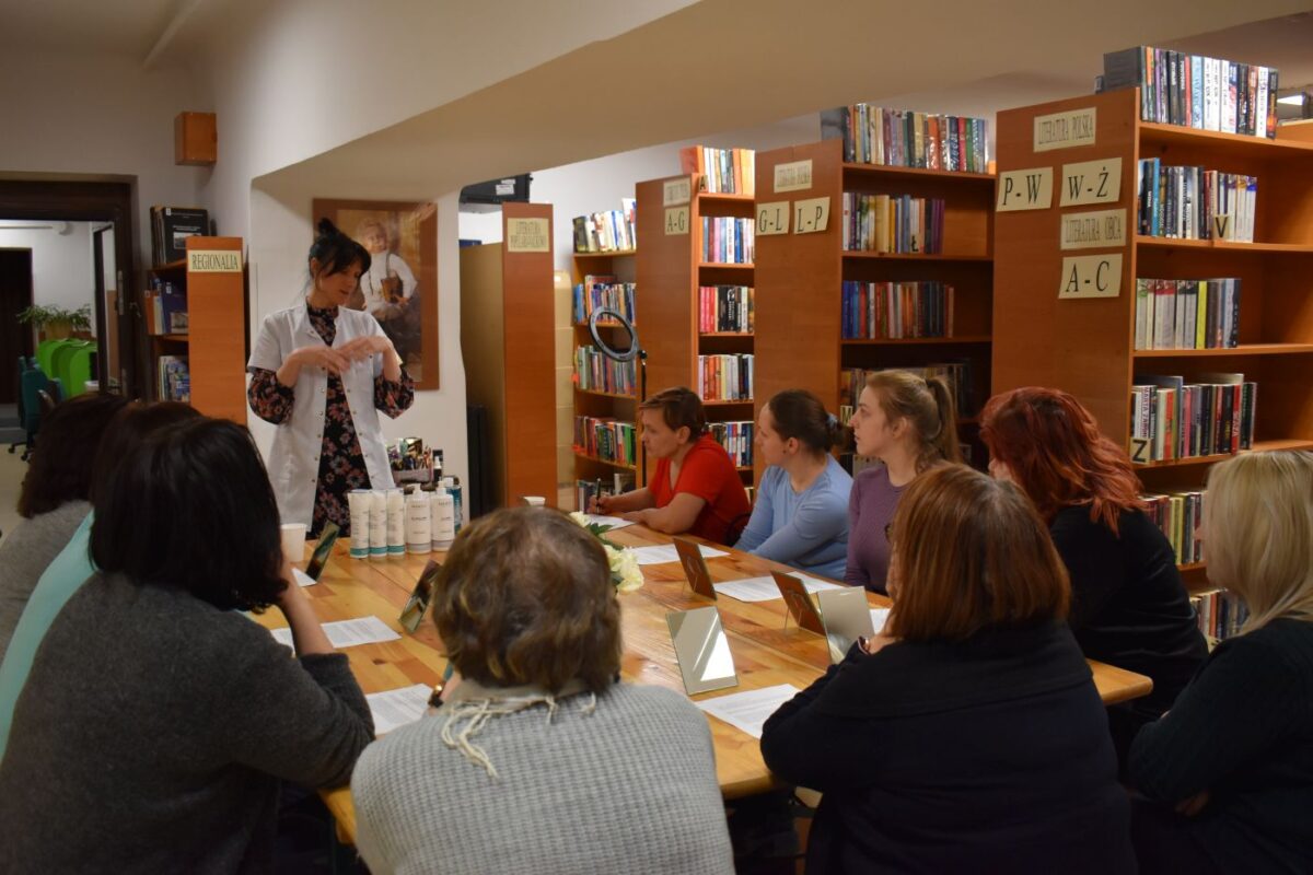 Stojąca kobieta w białym fartuchu mówi do pań siedzących przy stole, na którym stoją lusterka i leżą białe kartki papieru. W tle regały z książkami