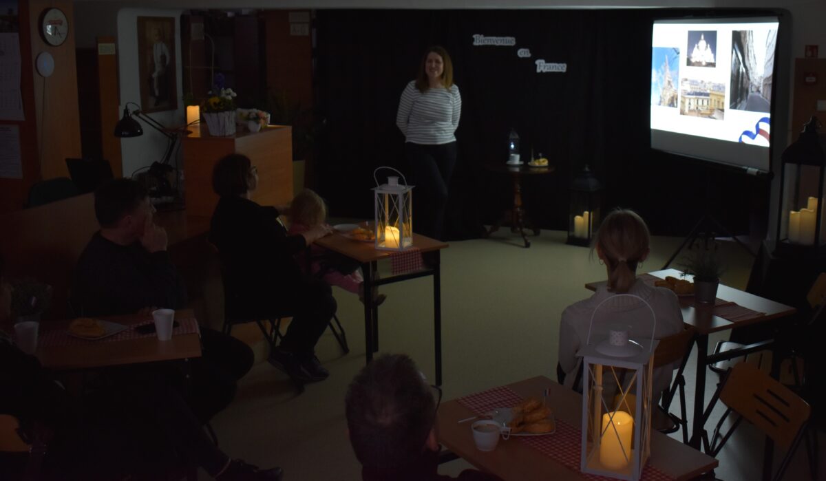 Kobieta stoi i patrzy na jasny ekran, przed nią stoją lampiony i stolik. W tle czarna kotara