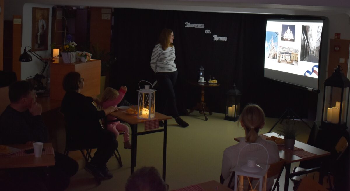 Przy stolikach na których stoją lampiony siedzą kobiety i mężczyźni. W oddali stoi kobieta przy ekranie.