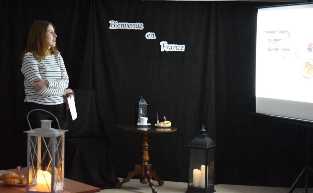 Kilka osób siedzi przy małych stolikach na których stoją lampiony na środku sali stoi kobieta w jasnej bluzce i patrzy na ekran. W tle czarna kotara