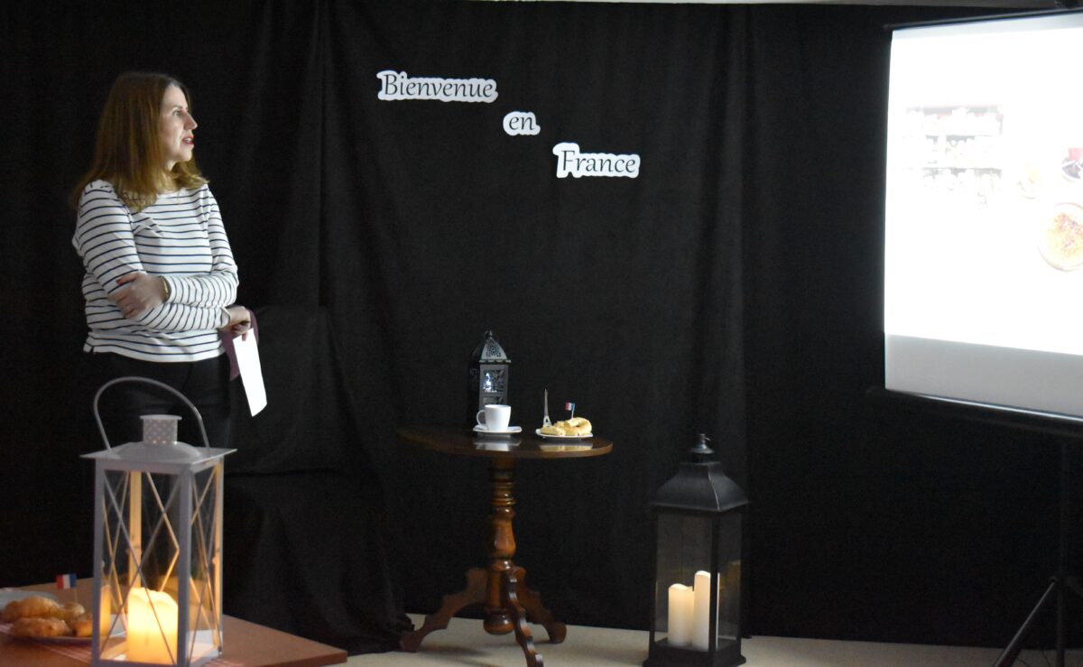 Kilka osób siedzi przy małych stolikach na których stoją lampiony na środku sali stoi kobieta w jasnej bluzce i patrzy na ekran. W tle czarna kotara