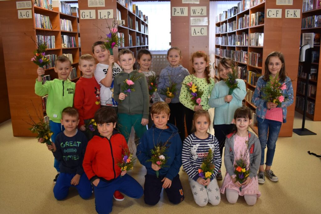 Grupka dzieci stoi na tle regałów bibliotecznych z książkami w reku trzymają zrobione przez siebie kolorowe palmy wielkanocne.