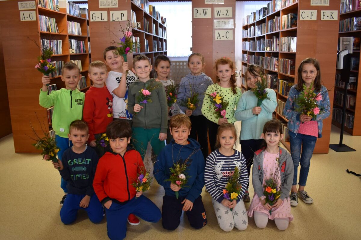 Grupka dzieci stoi na tle regałów bibliotecznych z książkami w reku trzymają zrobione przez siebie kolorowe palmy wielkanocne.