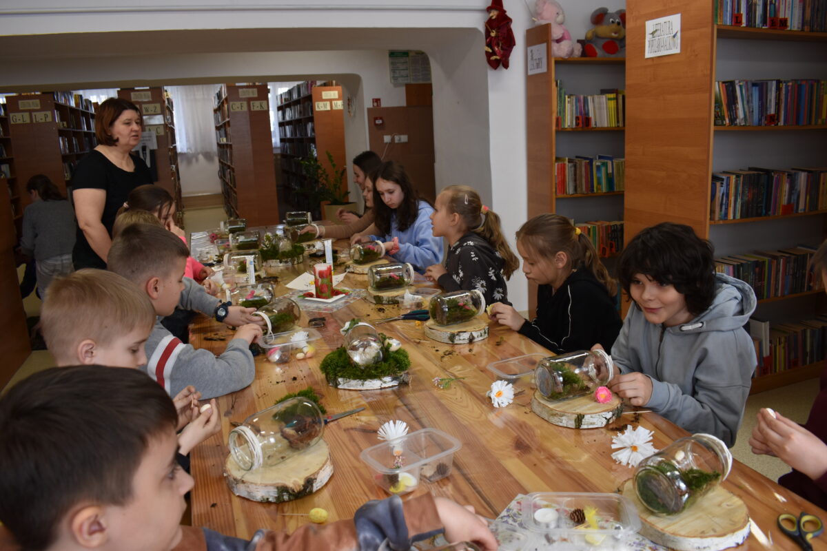 W sali bibliotecznej siedzą dzieci przy stole, przed nimi stoją słoiki w których robią dekoracje świąteczne. W tle stoi kobieta mówi do dzieci z nią regały z książkami