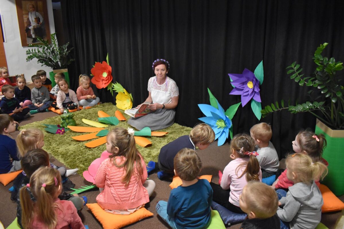 Grupa dzieci stoi a część siedzi na dywanie, w ręku trzymają zielone i czerwone tasiemki. W tle czarna kotara