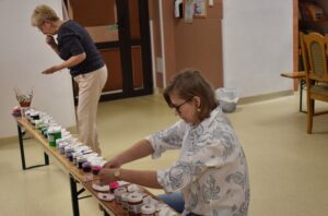 Jedna kobieta stoi , druga kuca przed ławką na której stoją dużo pojemniczków z kolorowymi farbami