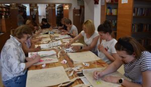 Przy drewnianym stole siedzą kobiety i malują na płóciennych torbach. W tle regały z książkami.