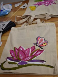 Na stole leży płócienna torba na której namalowana jest lilia w kolorach różu i fioletu z zieloną gałązką i kolorowym motylem