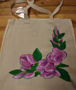 Na drewnianym stole leży torba z uszami na której namalowane są różowe kwiaty z zielonymi listkami.