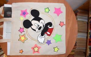 Torba płócienna na której namalowana jest Myszka Miki w kółku i wkoło kolorowe gwiazdki. Torba lezy na drewnianym stoliku obok stoją książki