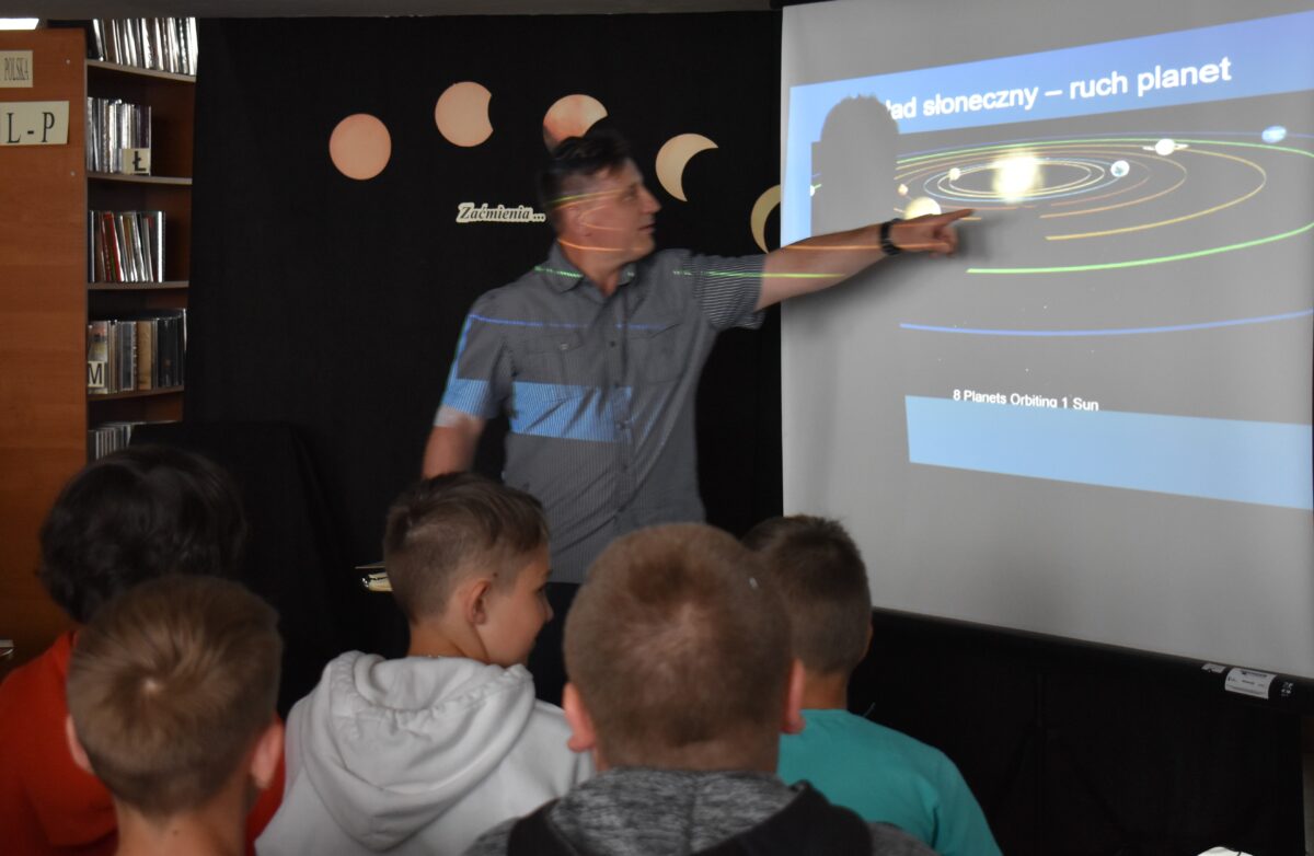 Mężczyzna pokazuje dzieciom ekranie układ słoneczny, pięciu chłopców mu się przygląda. W tle regały z książkami i czarna kotara na której pokazane jest zaćmienie słońca.
