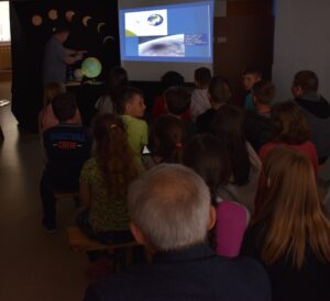 W ciemnej sali bibliotecznej dzieci siedzą na ławeczkach i oglądają slajdy w oddali stoi mężczyzna i wskazuje na globus