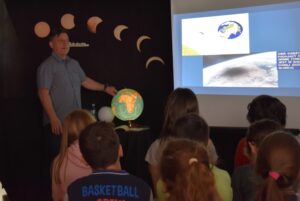 Stojący mężczyzna opiera rękę o podświetlony globus, przyglądają mu się dzieci. W tle ekran, czarna kotara na której pokazane jest zaćmienie słońca