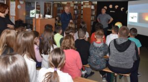 Grupa dzieci siedzących na ławkach w sali bibliotecznej, przed nimi stoi dwóch mężczyzn i kobieta w tle regały z książkami.