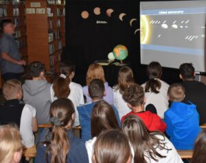 Przed ekranem siedzą dzieci i stoi mężczyzna na którym widać układ słoneczny, w tle regały z książkami