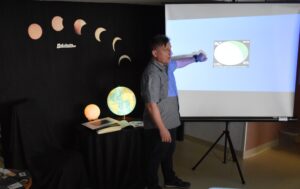 Mężczyzna stoi przed ekranem i coś pokazuje. W tle czarna kotara na której widać jak wygląda zaćmienie słońca, poniżej stoi podświetlony globus i księżyc, obok leżą książki i zdjęcia.