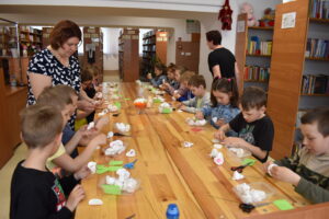 Dzieci siedzą przy stole i robią kwiatki w tle regały z książkami.