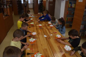 Przy drewnianym stole siedzą dzieci i robią bukiety dla swoich mam. W tle regały biblioteczne z książkami