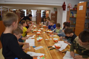 W sali bibliotecznej gdzie jest dużo regałów z książkami przy długim stole siedzą dzieci i robią laurki na Dzień Matki.
