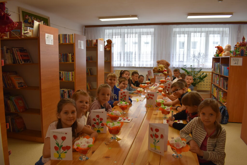 Uśmiechnięte dzieci siedzą przy stole w ręku trzymają laurki i bukiety dla swoich Mam. W tle regały z kolorowymi książkami