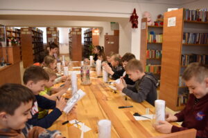 Gromadka dzieci siedzi przy stole i w reku trzymają szklane wazony na których malują. W tle regały z książkami