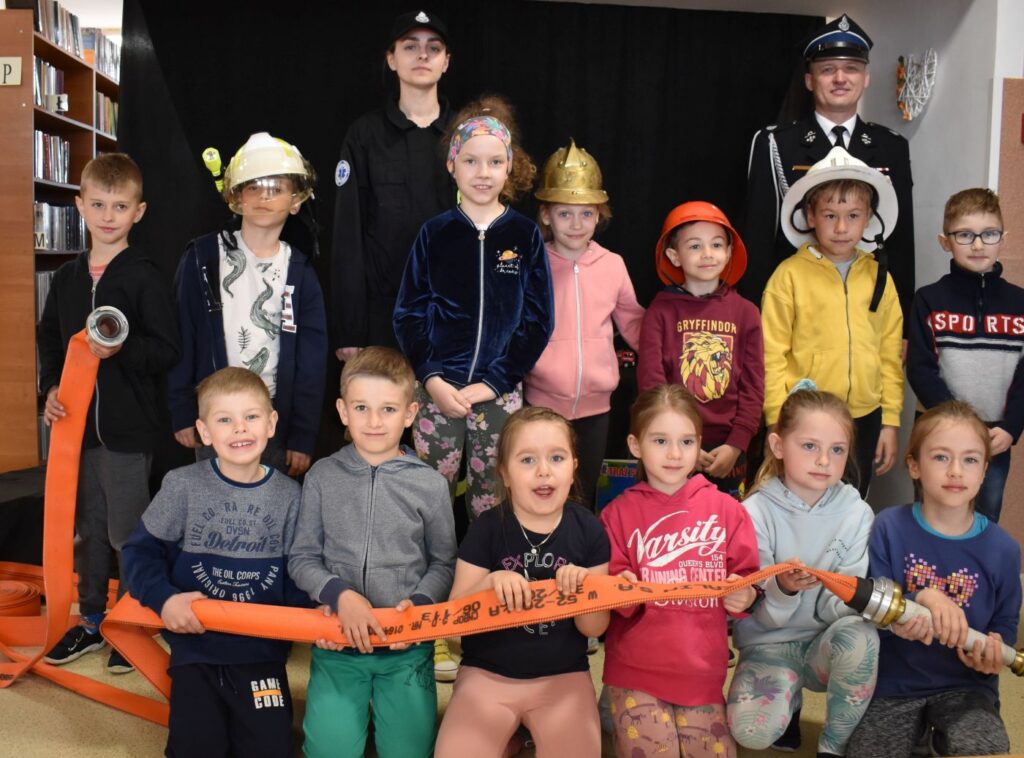 Uśmiechnięta grupka dzieci w kaskach strażackich, trzyma wąż strażacki za nimi stoją strażacy w mundurach.