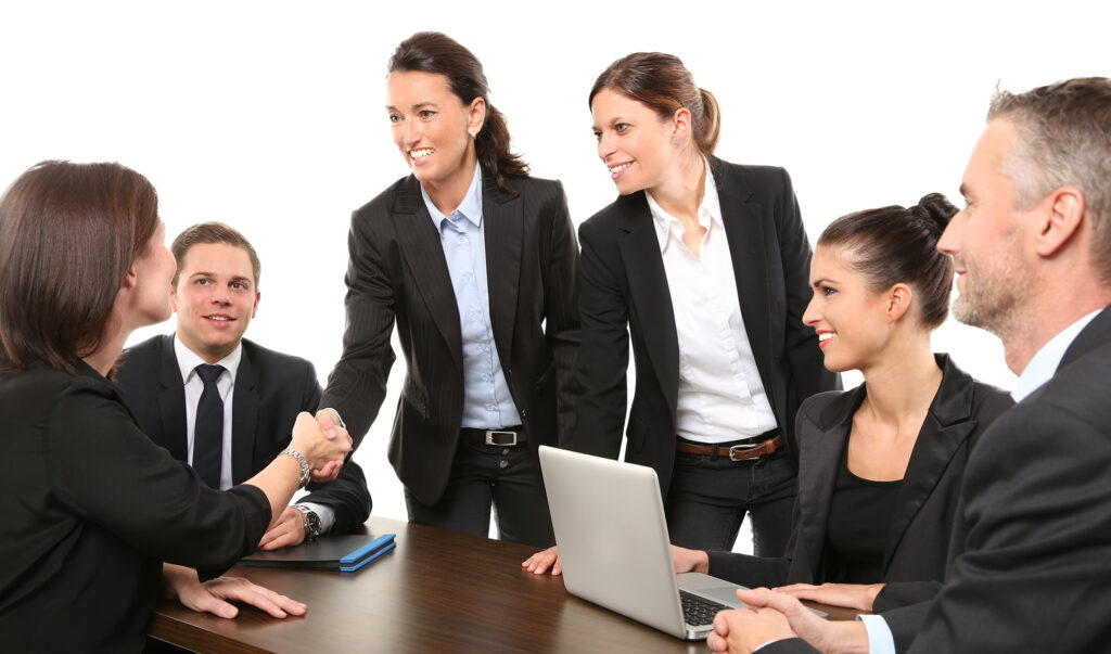 Kilka osób przy stoliku, ubranych w eleganckie czarnobiałe stroje. Jedna z kobiet uściska dłoń z siedzącą kobietą.