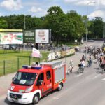 Czerwony wóz strażacki za nim dużo rowerzystów na skrzyżowaniu w tle park